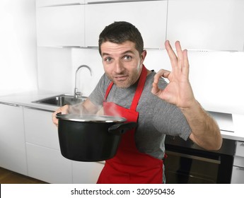 Young Successful Man At Home Kitchen In Red Cook Apron Holding Pot Delighted With The Good Smell And Taste Of His Dish Proud , Satisfied And Happy In Cooking Success Concept