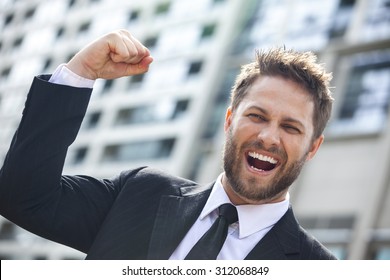 A young successful man, male executive businessman arms raised celebrating cheering shouting in front of a high rise office block in a modern city