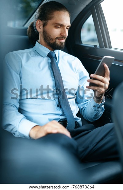 Young Successful Man Working On Phone Sitting In\
Car. Portrait Of Handsome Smiling Businessman In Formal Clothes\
Using Phone Riding To Work On Back Seat Of Luxury Business Car. \
High Quality Image.