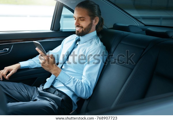 Young Successful Man Working On Phone Sitting In
Car. Portrait Of Handsome Smiling Businessman In Formal Clothes
Using Phone Riding To Work On Back Seat Of Luxury Business Car. 
High Quality Image.