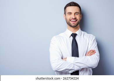 Young and successful. Portrait of handsome young man in shirt and tie keeping arms crossed and smiling while standing against grey background - Powered by Shutterstock