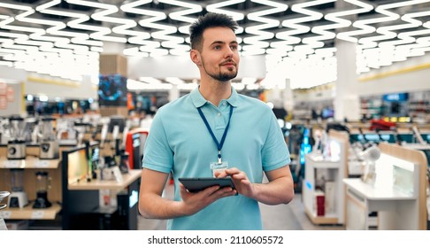 A Young Successful Sales Assistant In Uniform With A Tablet In His Hands Against The Background Of The Interior Of The Hall In A Home Appliances And Electronics Store.