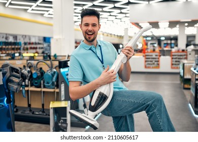 A Young Successful Sales Assistant In Uniform Having Fun Playing A Vacuum Cleaner Like A Guitar Against The Background Of The Interior Of A Hall In A Home Appliances And Electronics Store.