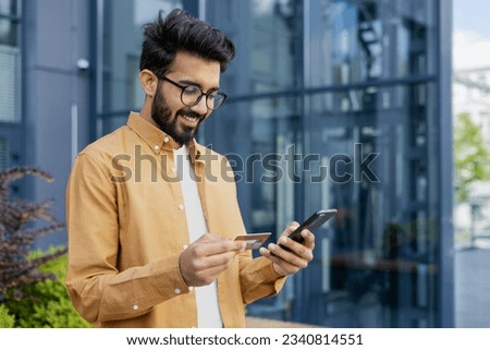 Young successful satisfied hispanic man smiling outside office building, businessman using app on phone for online shopping in online store, man holding bank credit card and smartphone.