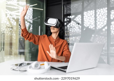 Young successful woman inside the office using vr glasses, virtual reality simulator cross charts and documents online, latin american woman using laptop at work. - Powered by Shutterstock