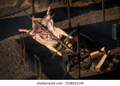A young suckling pig on a skewer. Whole roasted pig on a rotating steel spit with fire and smoke. Traditional grill of Serbia, in Balkans, countryside.