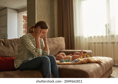 Young Suffering Woman Sitting On Couch, Holding Head In Hands, Little Crying Baby Lying Near. Highly Sensitive Mother Suffering From Chronic Headache Caused By Instant Baby Crying.