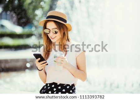 Young summer beauty woman with phone chating or surf in internet drinks mojito fruit cocktail against fountain in the street