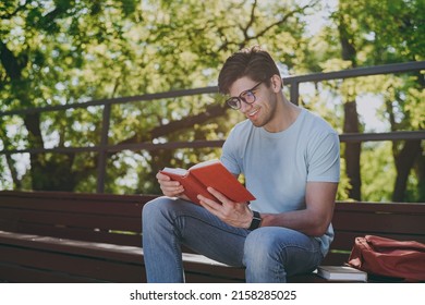 Young sunlit student man 20s wearing blue t-shirt eyeglasses backpack read book sitting on bench walking rest relax in sunshine spring green city park outdoors on nature. Education high school concept