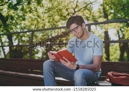 Young sunlit student man 20s wearing blue t-shirt eyeglasses backpack read book sitting on bench walking rest relax in sunshine spring green city park outdoors on nature. Education high school concept