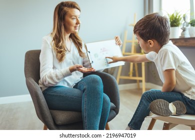 Young Supported Adoption Counselor Showing A Drawing Of House To Sad Pensive Little Boy Waiting For New Loving Family. Young Child Psychologist Working With Little Boy