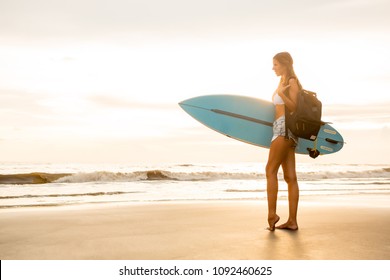 Young surfer girl in sexy white bikini with surf board and travel backpack pose on sunset beach. Modern family lifestyle, people water sport adventure camp and extreme swim on summer vacation