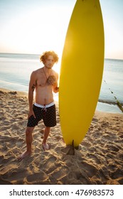 Young surfer holding surf board while standing at the beach at sunset