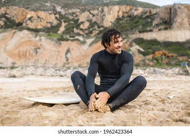 Young surfer man wearing diving suit sitting on the sand next to his surfboard and resting after surfing. Smiling and talking with somebody - Shutterstock ID 1962423364