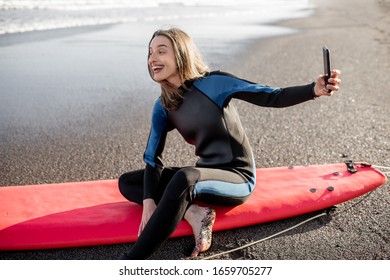 Young Surfer In Wetsuit Making Self Portrait On Phone While Sitting With Surfboard On The Beach. Water Sport And Active Lifestyle Concept