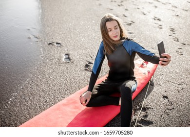 Young Surfer In Wetsuit Making Self Portrait On Phone While Sitting With Surfboard On The Beach. Water Sport And Active Lifestyle Concept