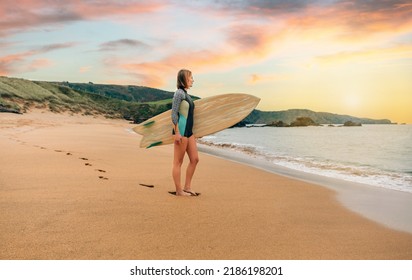 Young Surfer Woman With Wetsuit Carrying Surfboard Looking To The Sea At The Beach