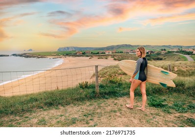 Young surfer woman with wetsuit and surfboard looking at the beach from the shore - Powered by Shutterstock