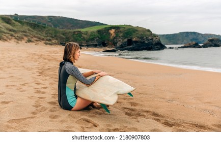 Young Surfer Woman With Wetsuit And Surfboard Sitting On The Sand Looking At The Sea