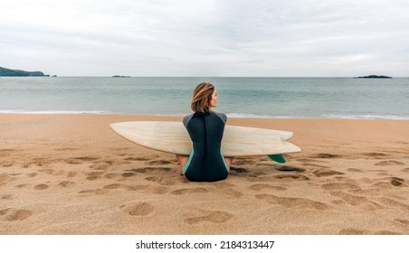 Young surfer woman with wetsuit and surfboard sitting on the sand looking aside on the beach