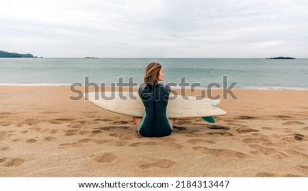Young surfer woman with wetsuit and surfboard sitting on the sand looking aside on the beach 商業照片 © 