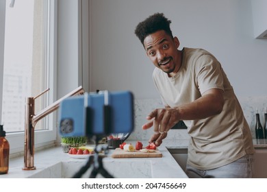 Young Surprised African American Man In Casual Clothes Prepare Sweet Breakfast Cut Fruit Watch Series On Mobile Cell Phone Cooking Food In Indoor Kitchen At Home Alone Healthy Diet Lifestyle Concept.