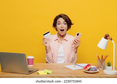 Young surprised amazed employee business woman in casual shirt sit work at office desk use mobile cell phone credit card book tour isolated on plain yellow color background Achievement career concept