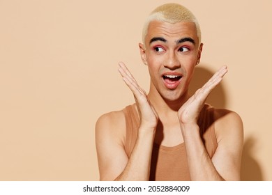 Young surprised amazed fun happy blond latin american gay man 20s with make up in beige tank shirt look aside isolated on plain light ocher background studio portrait People lgbt lifestyle concept.