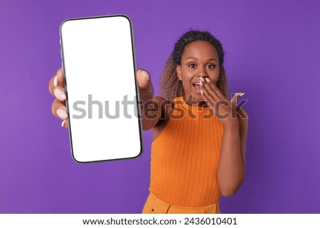 Young surprised beautiful African American woman shows phone with blank screen and experiencing positive shock after receiving sms newsletter with lucrative offer stands in purple studio.