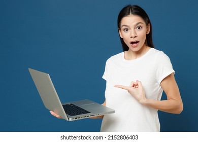 Young surprised copywriter freelancer student positive latin woman 20s wear white casual basic t-shirt point index finger on laptop pc computer isolated on dark blue color background studio portrait