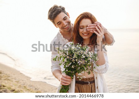Young surprised couple family man woman in white clothes rest relax together boyfriend meet girlfriend close eyes gift give bouquet flowers at sunrise over sea sand beach outdoor seaside in summer day