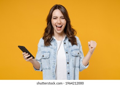 Young surprised excited brunette fun woman in stylish casual denim shirt white t-shirt hold mobile cell phone do winner gesture clench fist celebrating isolated on yellow background studio portrait.