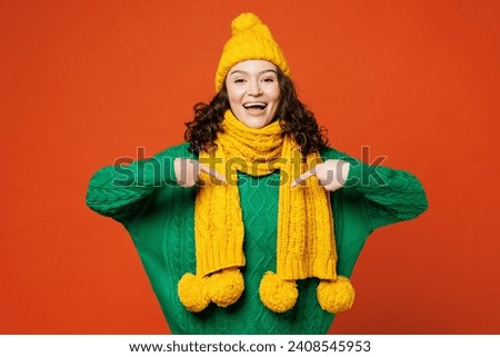 Young surprised excited impressed happy woman she wear green knitted sweater yellow hat scarf point index finger on herself isolated on plain orange red background studio portrait. Lifestyle concept