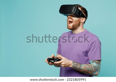 Young surprised excited man he wears purple t-shirt hold in hand play pc game with joystick console watching in vr headset pc gadget isolated on plain pastel light blue cyan background studio portrait