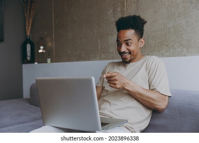 Young surprised fun african american man in casual beige t-shirt sweatpants sitting on grey sofa indoors apartment point index finger on laptop pc computer waving hand resting on weekends stay home