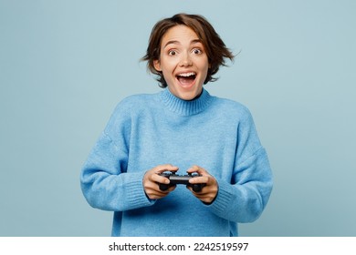 Young surprised fun caucasian woman wear knitted sweater hold in hand play pc game with joystick console isolated on plain pastel light blue cyan background studio portrait. People lifestyle concept