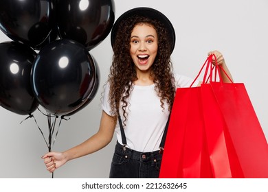 Young Surprised Fun Excited Woman Wear T-shirt Hat Hold In Hand Red Paper Package Bags After Shopping Balloons Isolated On Plain Solid White Color Background Studio. Black Friday Sale Buy Day Concept