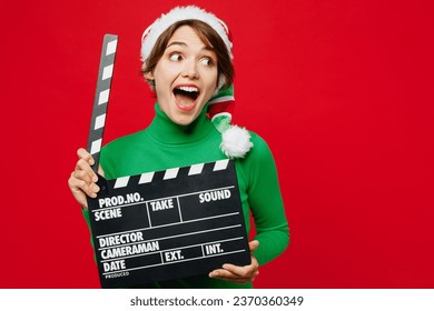 Young surprised fun woman wear green turtleneck Santa hat posing hold classic black film making clapperboard look aside isolated on plain red background. Happy New Year 2024 Christmas holiday concept