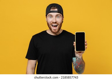 Young surprised happy bearded tattooed man 20s he wears casual black t-shirt cap hold in hand use mobile cell phone with blank screen workspace area isolated on plain yellow wall background studio.