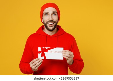 Young surprised happy fun man wears red hoody hat look camera hold gift certificate coupon voucher card for store isolated on plain yellow color background studio portrait. People lifestyle concept