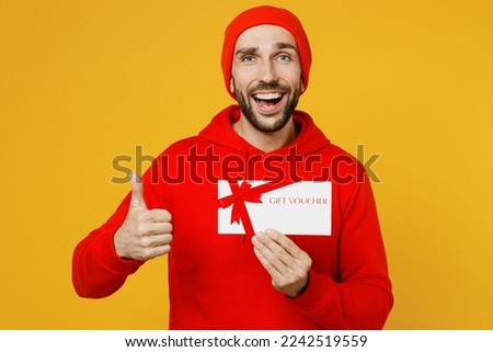 Young surprised happy fun man wearing red hoody hat look camera hold gift certificate coupon voucher card for store show thumb up isolated on plain yellow color background. People lifestyle concept