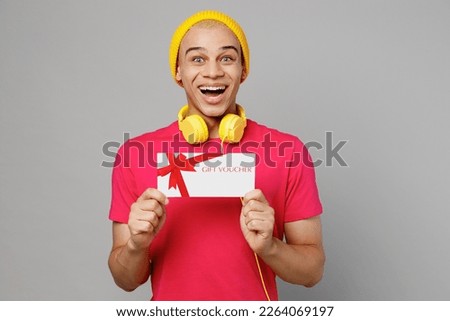 Young surprised happy fun man of African American ethnicity 20s he wear pink t-shirt yellow hat headphones hold gift certificate coupon voucher card for store isolated on plain grey background studio