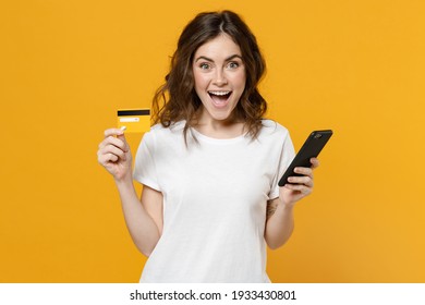 Young Surprised Happy Positive Caucasian Student Woman 20s In White Basic Casual T-shirt Look Camera Hold Mobile Cell Phone Credit Bank Card Isolated On Yellow Orange Color Background Studio Portrait