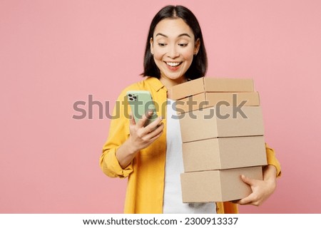Young surprised happy woman of Asian ethnicity wears yellow shirt white t-shirt hold stack cardboard blank boxes use mobile cell phone isolated on plain pastel light pink background studio portrait