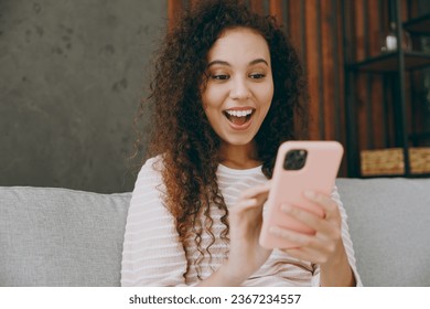 Young surprised happy woman wear casual clothes sits on grey sofa couch hold in hand use mobile cell phone stay at home hotel flat rest relax spend free spare time in living room indoor Lounge concept - Shutterstock ID 2367234557
