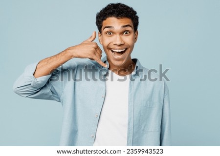 Young surprised man of African American ethnicity wear shirt white t-shirt casual clothes doing phone gesture like says call me back isolated on plain pastel light blue cyan background studio portrait