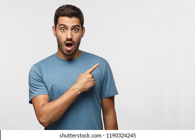 Young surprised man in blue t-shirt pointing right with his finger, shouting WOW, isolated on gray background with copy space fot your text. Shock content