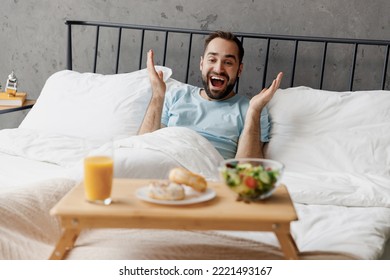 Young Surprised Man Wear Blue T-shirt Lying In Bed Spread Hands Look At Breakfast Table Hands Rest Relax Spend Time In Bedroom Lounge Home In Own Room House Wake Up Dream. Good Mood Bedtime Concept