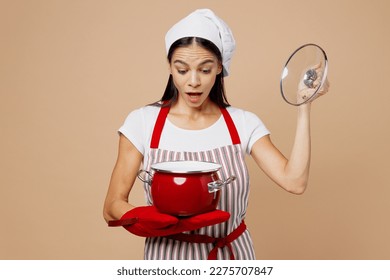 Young surprised shocked housewife housekeeper chef baker latin woman wear striped apron toque hat hold in hand red pot pan lift lid isolated on plain pastel light beige background. Cook food concept - Powered by Shutterstock