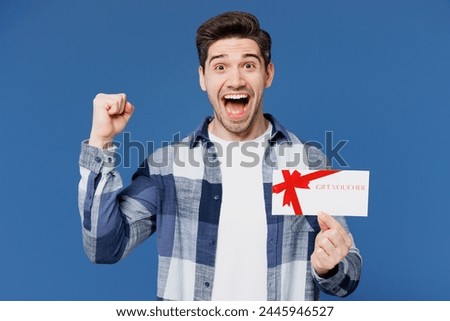 Young surprised shocked man he wears shirt white t-shirt casual clothes hold gift certificate coupon voucher card for store isolated on plain blue cyan background studio portrait. Lifestyle concept
